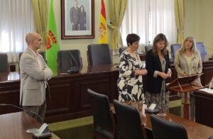 Salón de Rectorado con muebles de madera. La Dra. Gracia Merino se encuentra junto a la Rectora y la Secretaria General, tres mujeres de mediana edad, frente a un atril. A la izquierda se encuentra el Director del IBIOMED, José Antonio Molina, un hombre en traje.